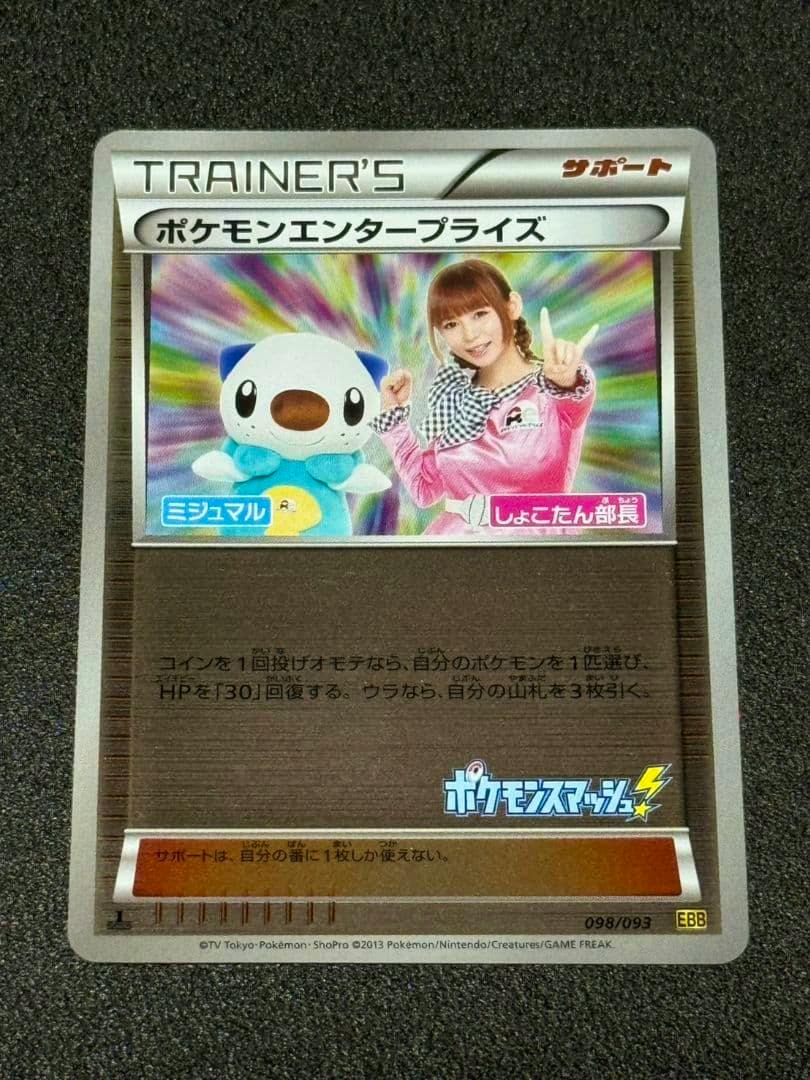 Photograph of the Pokémon trading card (BW concept pack “EX Battle Boost” mirror) showing a Trainer's support card with an Oshawott plush and a woman in pink against a rainbow background.