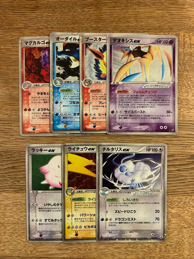 Seven Pokémon trading cards (ex cards) arranged on a wooden surface, showing holographic artwork including a visible Deoxys-ex card.