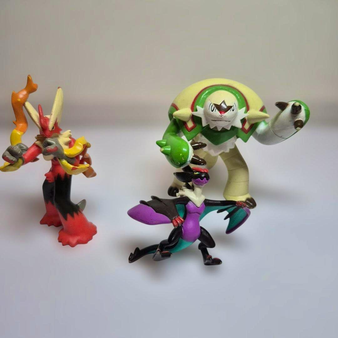Three Pokémon plastic figures on a white surface: a red-and-yellow bipedal figure, a purple-and-black winged figure, and a large green-and-cream armored figure.