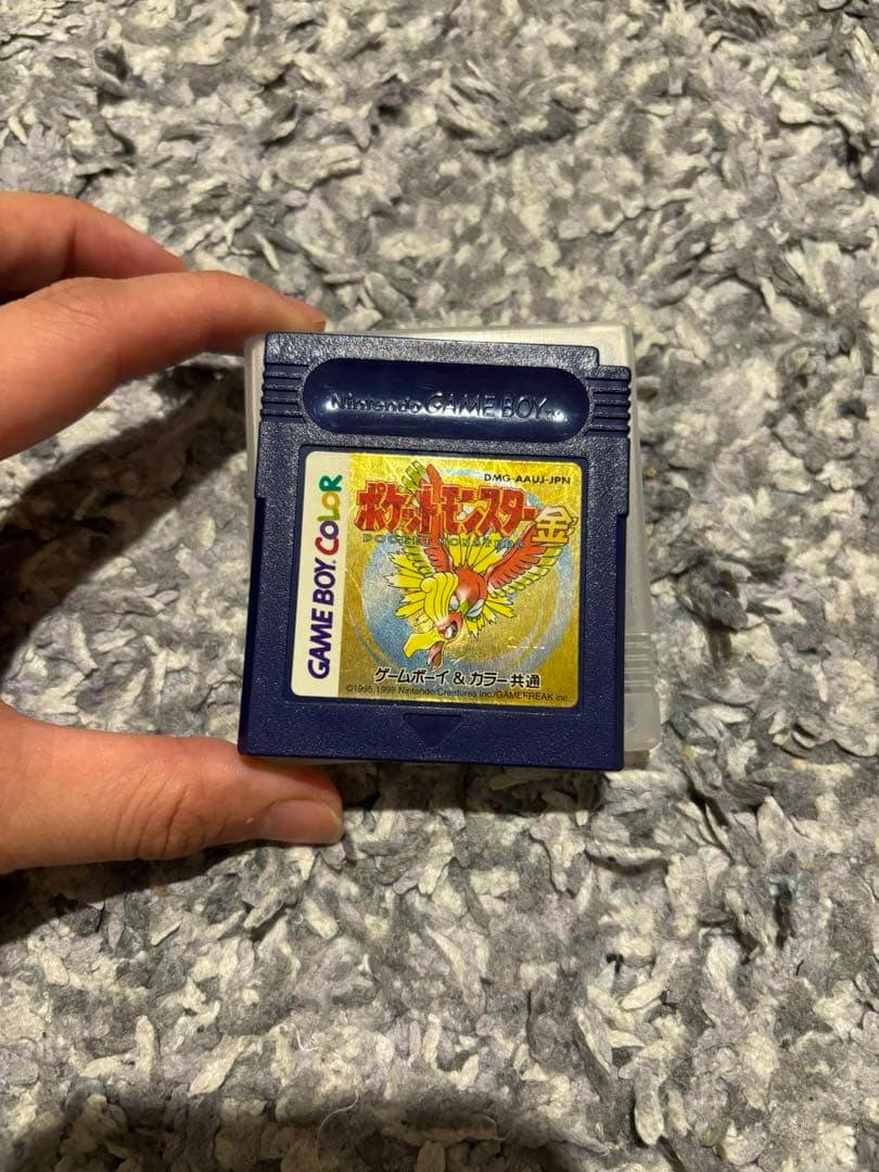 A hand holding the front of a Game Boy Color cartridge for ポケットモンスター金 (Pocket Monsters Gold) on a textured surface.