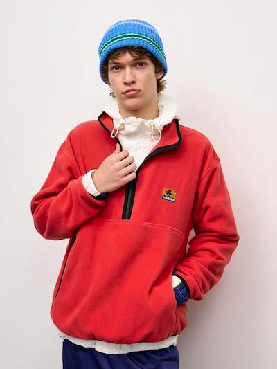 Male model wearing a red half-zip fleece pullover with a black zipper and a small Pokémon patch on the chest.