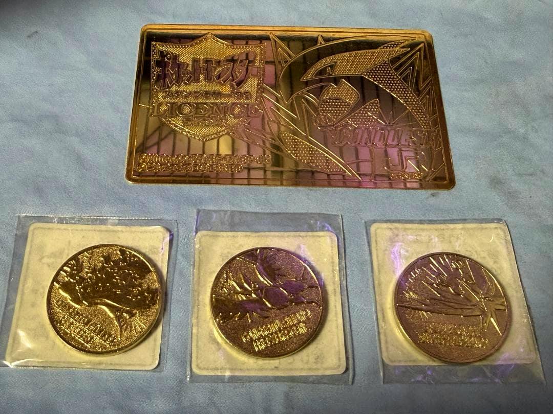 A gold-colored rectangular Pokémon JR West "Licence" plate shown above three round gold medals in clear plastic sleeves on a blue cloth.