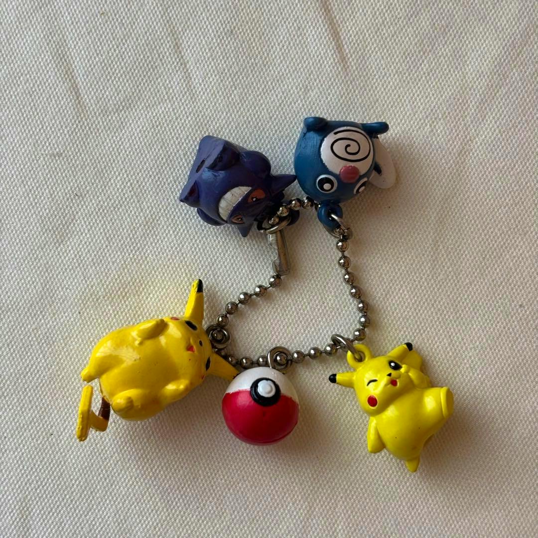 A plastic Pokémon keychain showing Pikachu, Gengar, Poliwag and a Poké Ball charms on a ball chain displayed on white fabric.