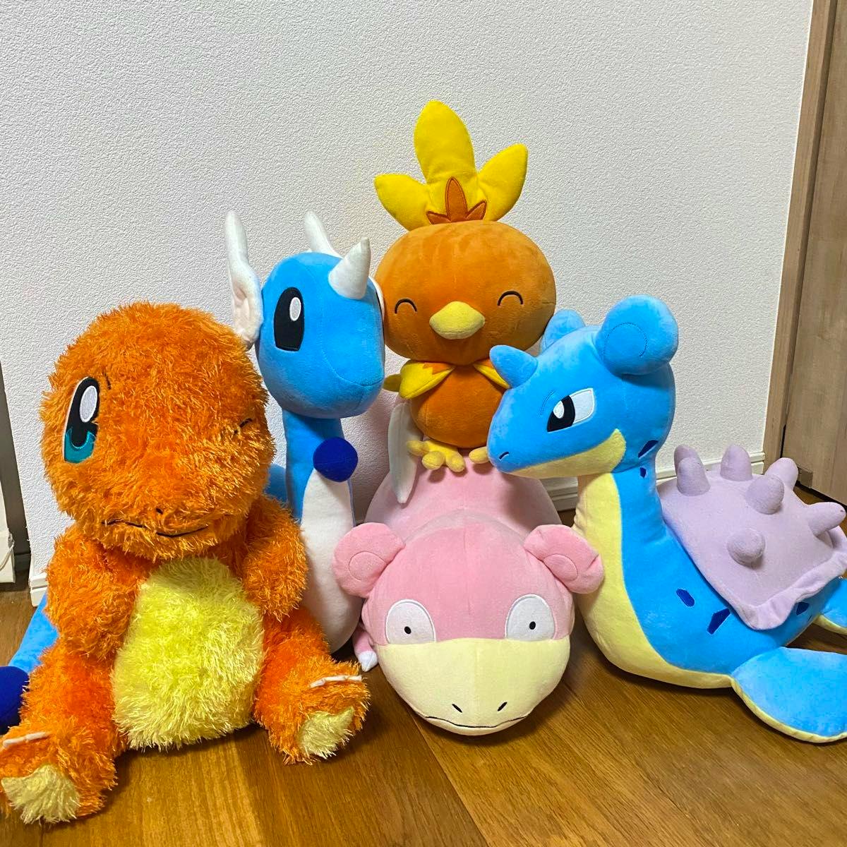 Several Pokémon plush toys including Charmander, Dragonair (Hakuryu), Slowpoke (Yadon), and Lapras arranged on a wooden floor against a white wall.