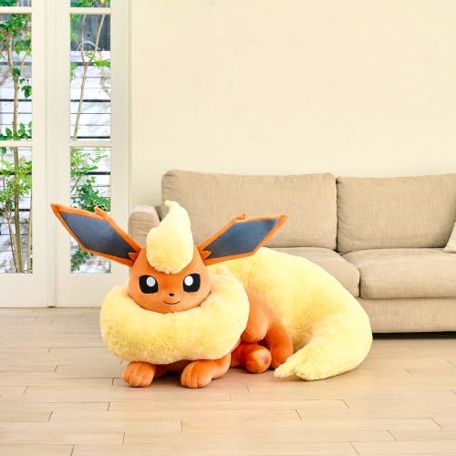 A life-size Flareon (Booster) lying plush toy displayed on a wooden floor in front of a beige sofa.