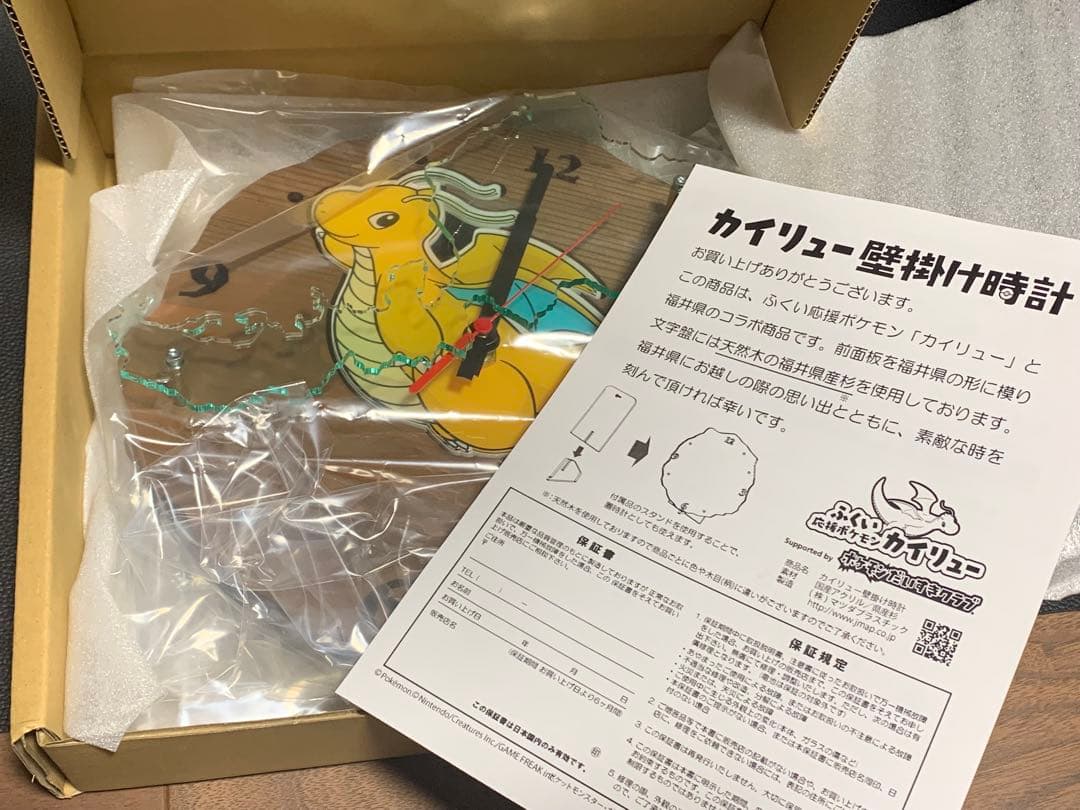 Opened box showing the Dragonite wall clock wrapped in plastic with the instruction sheet visible.