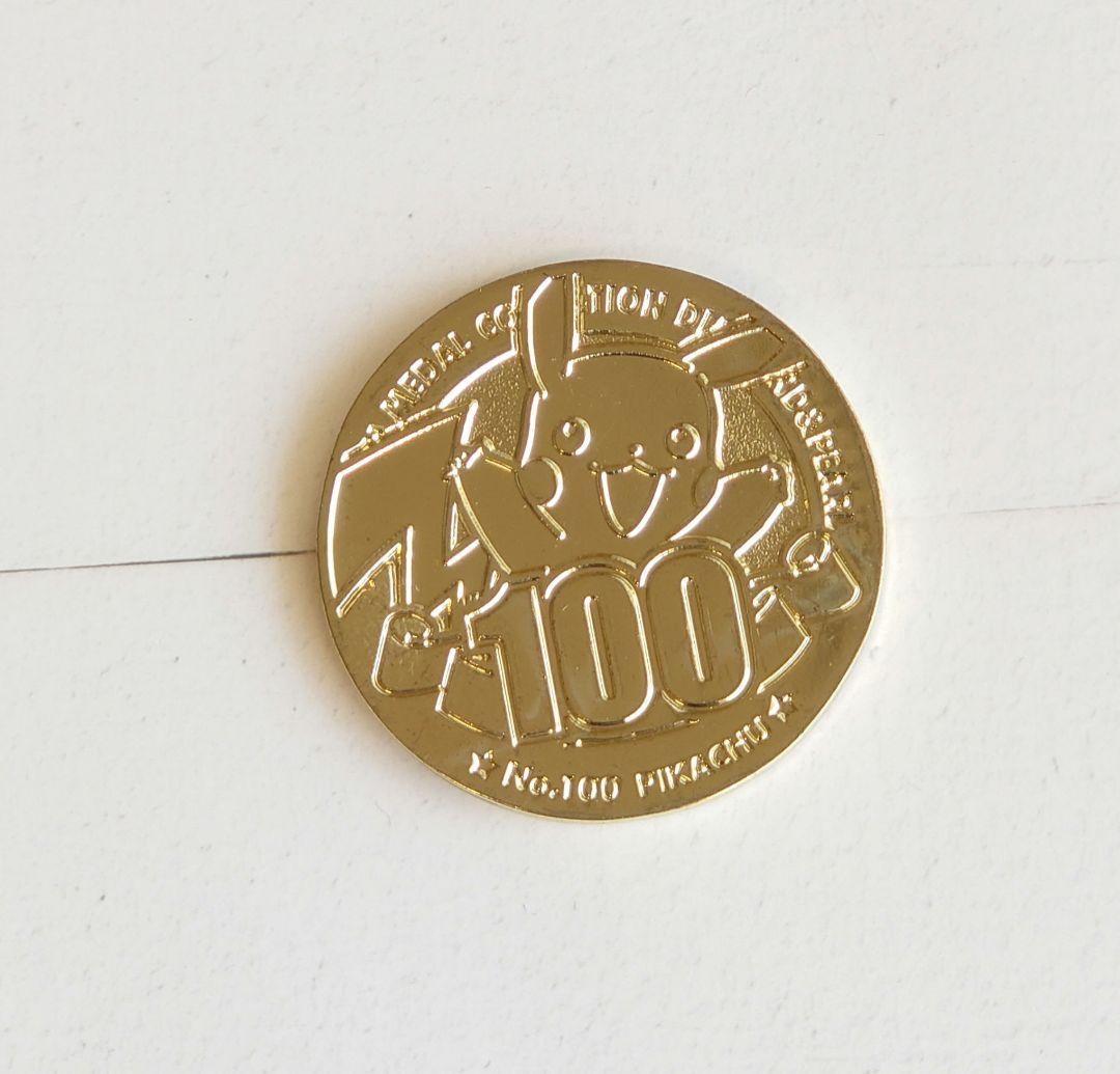 A gold-colored Pokémon commemorative medal face showing Pikachu and the number "100" on a white background.