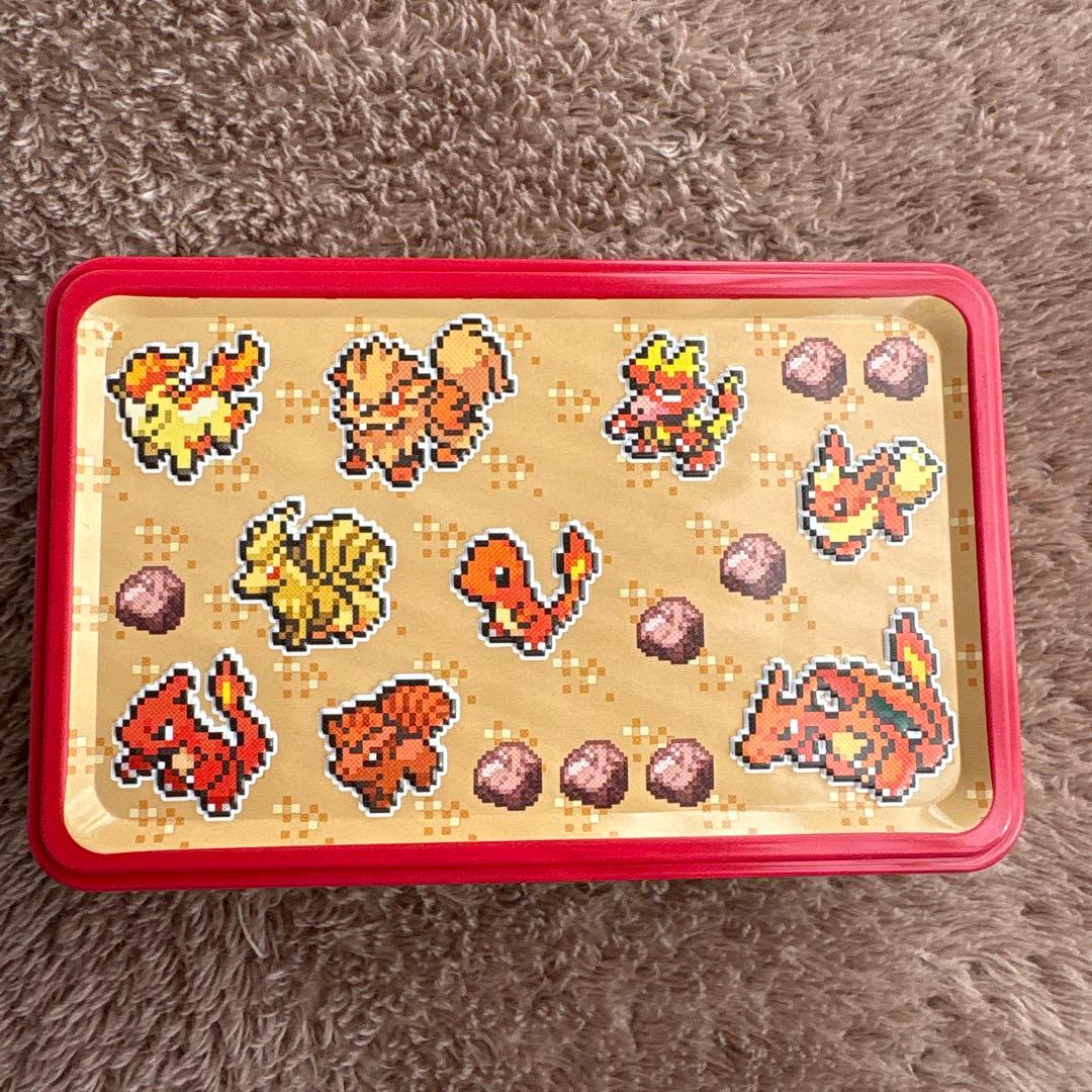 Rectangular red-lidded tin with pixel-art Pokémon characters printed on the lid, shown on a textured fabric surface.