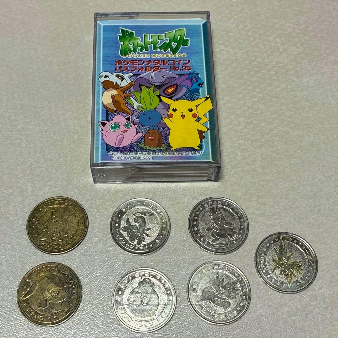 A Pokémon pass holder case labeled No.25 in a clear plastic box with seven metal Pokémon coins arranged below it.