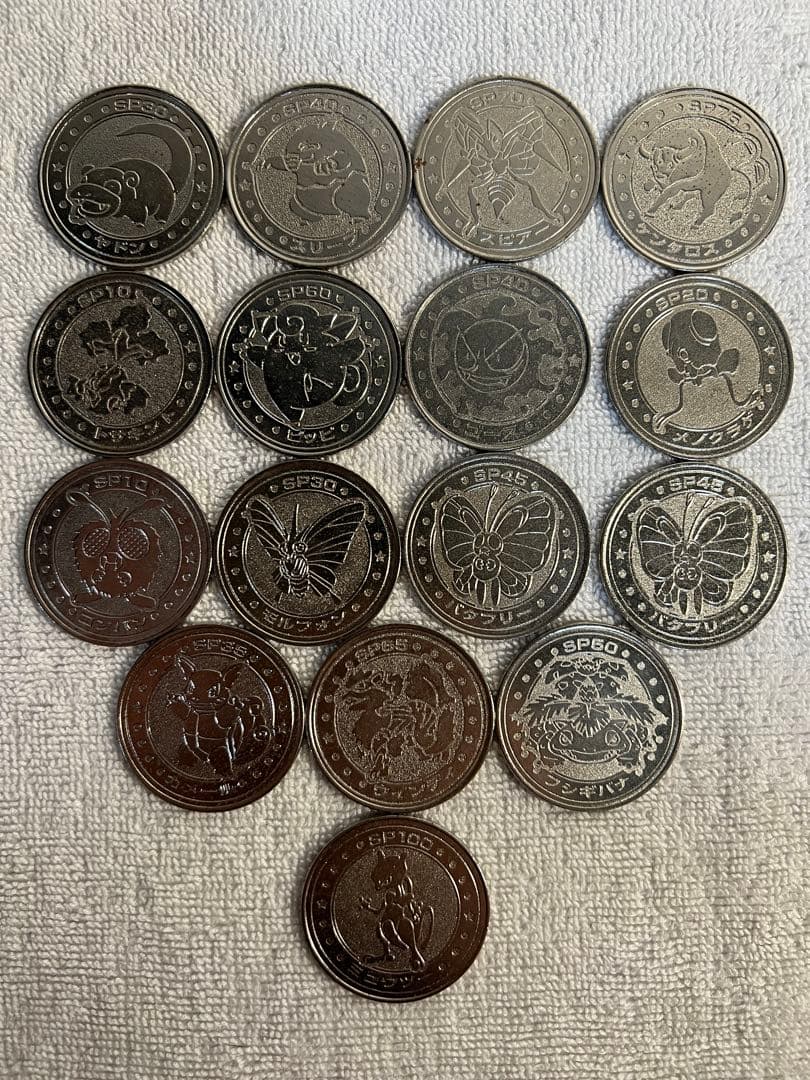 Sixteen Pokémon metal coins arranged on a white towel showing the coins' front faces with various Pokémon designs.