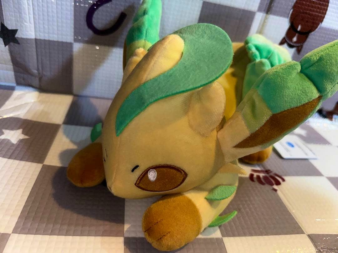 Front-angle photo of a Leafeon plush toy lying on a checkered mat, showing its face and green leaf details.