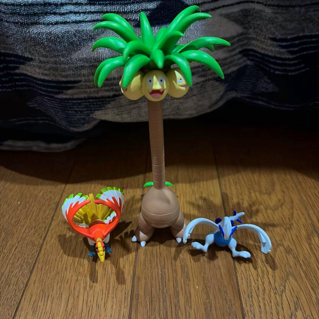 Three Pokémon Moncolle plastic figures — Alolan Exeggutor in the center with Ho-Oh (red) and Lugia (blue) on a wood floor.