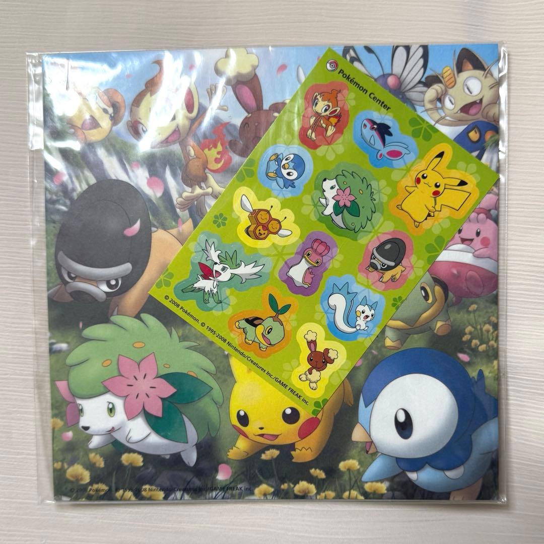 A sealed pack of Pokémon origami paper with a Pokémon sticker sheet (featuring Shaymin) placed on top.