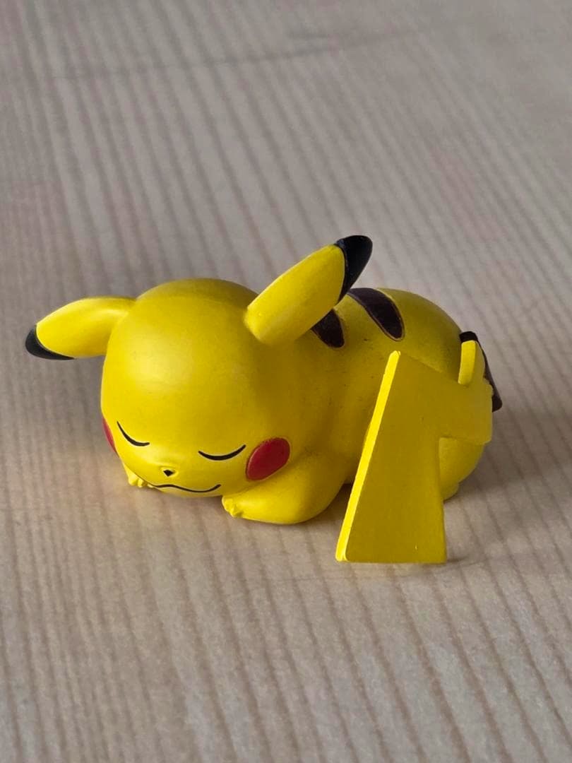 A yellow sleeping Pikachu plastic figure lying on its belly with a separate yellow tail-shaped stand on a wood-grain surface.
