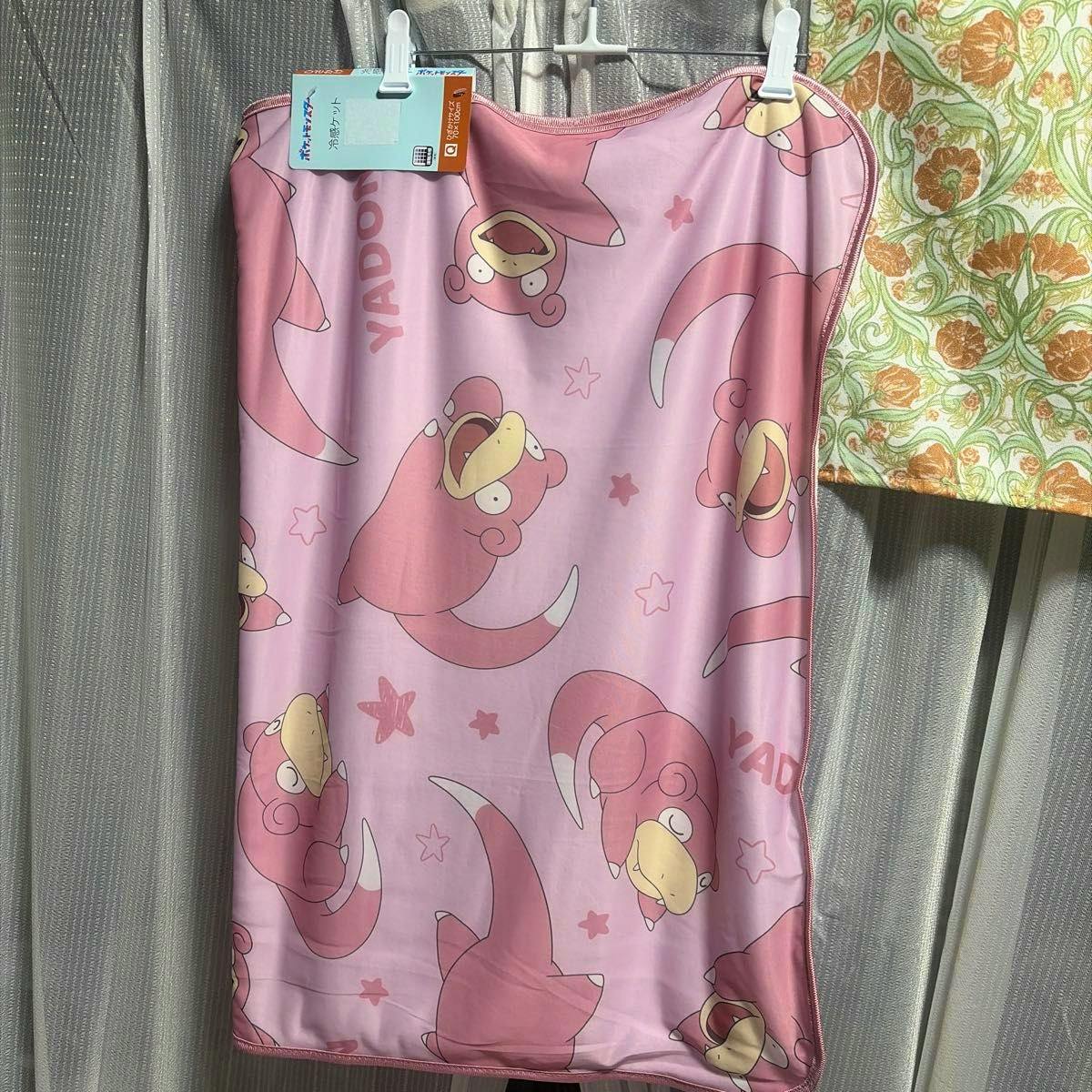 Pink Pokémon Slowpoke (ヤドン) blanket with repeated character print and attached tag, hung by clips against curtains.