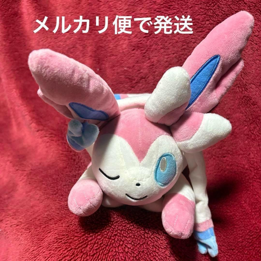 Front view of a Pokémon ニンフィア (Sylveon) pink and white plush toy lying on a red blanket with Japanese shipping text at the top.