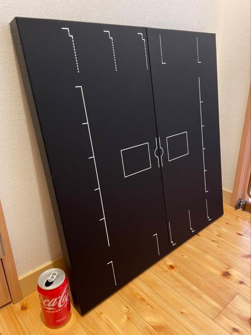 Black Pokémon card playmat with white printed layout markings leaning against a wall next to a Coca-Cola can for scale.