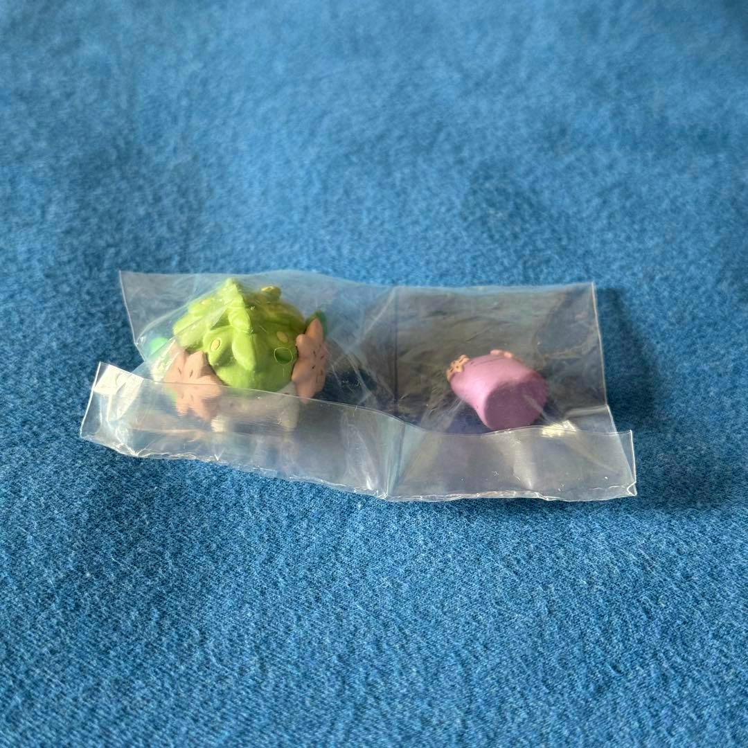 Two small Pokémon figure pieces (one green, one purple) sealed in clear plastic bags on a blue fabric surface.