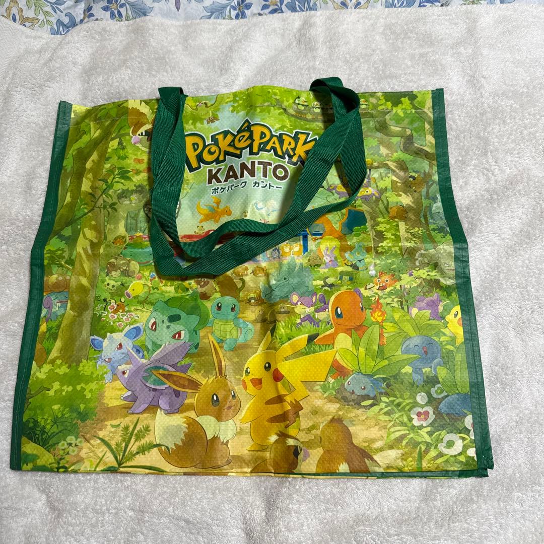 A green reusable tote bag printed with a PokePark KANTO forest scene featuring Pikachu, Eevee, Bulbasaur, Squirtle, Charmander and other Pokémon.