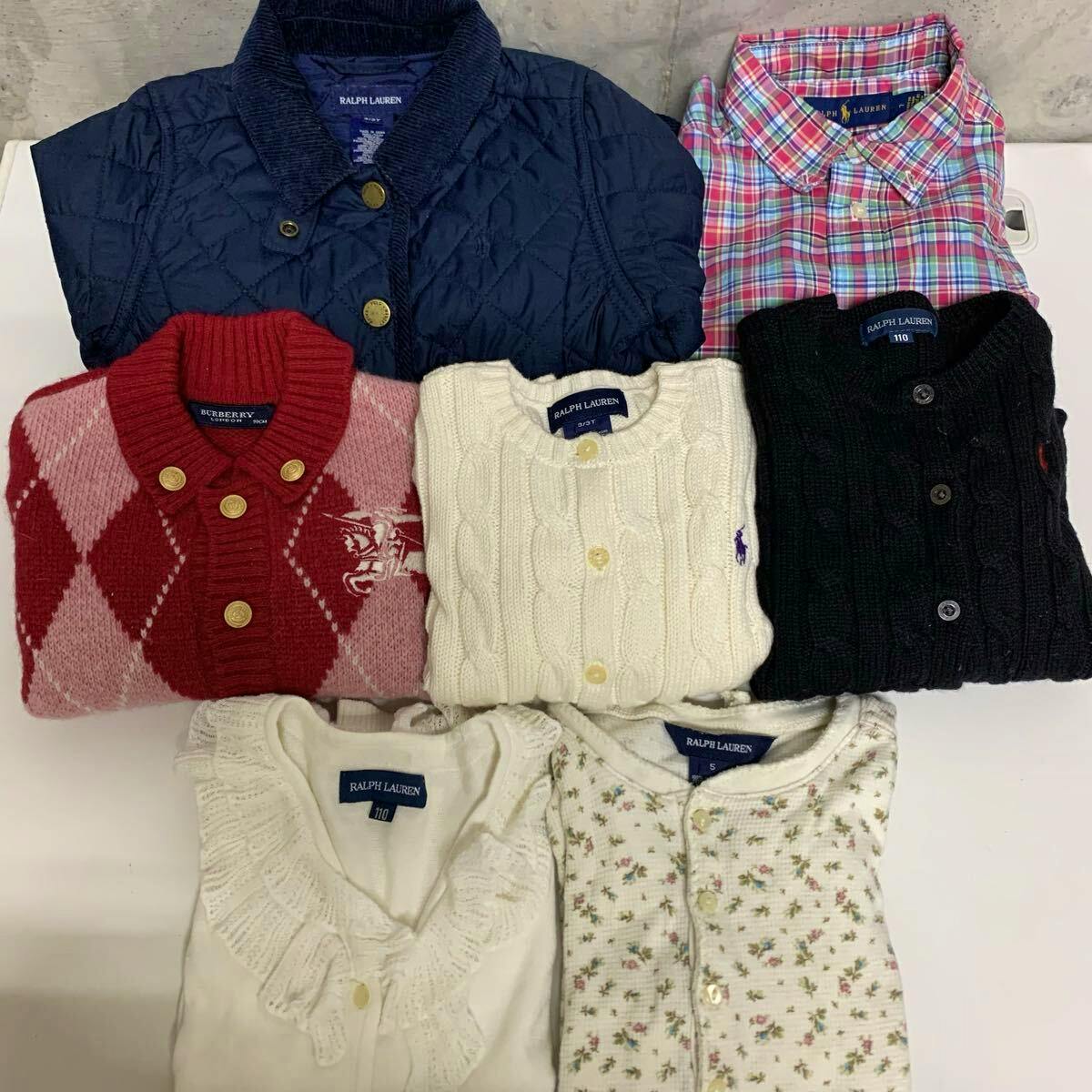 Top-down photo of folded kids sweaters and tops, including Ralph Lauren and Burberry items, arranged on a white surface.