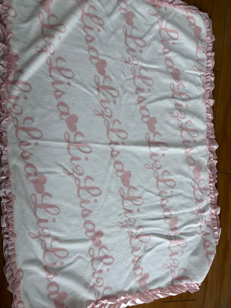 A white polyester blanket with a repeating pink script pattern and pink ruffled trim laid flat on a wooden floor.