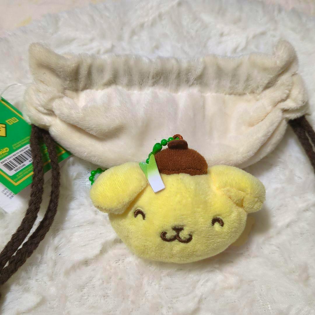 Yellow Pompompurin gyoza-shaped plush mascot pouch with a brown hat and green bead chain, shown in front of a cream drawstring pouch on a white furry background.
