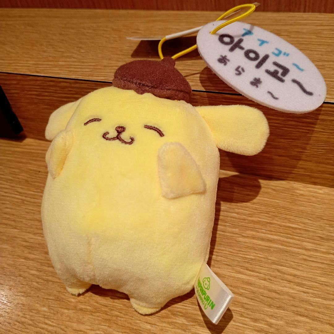 A Pompompurin yellow plush mascot with a brown beret and attached tag on a wooden surface.