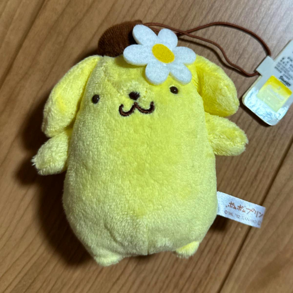 Yellow Pompompurin plush keychain mascot with a white felt flower on its head and an attached tag, shown on a wooden surface.