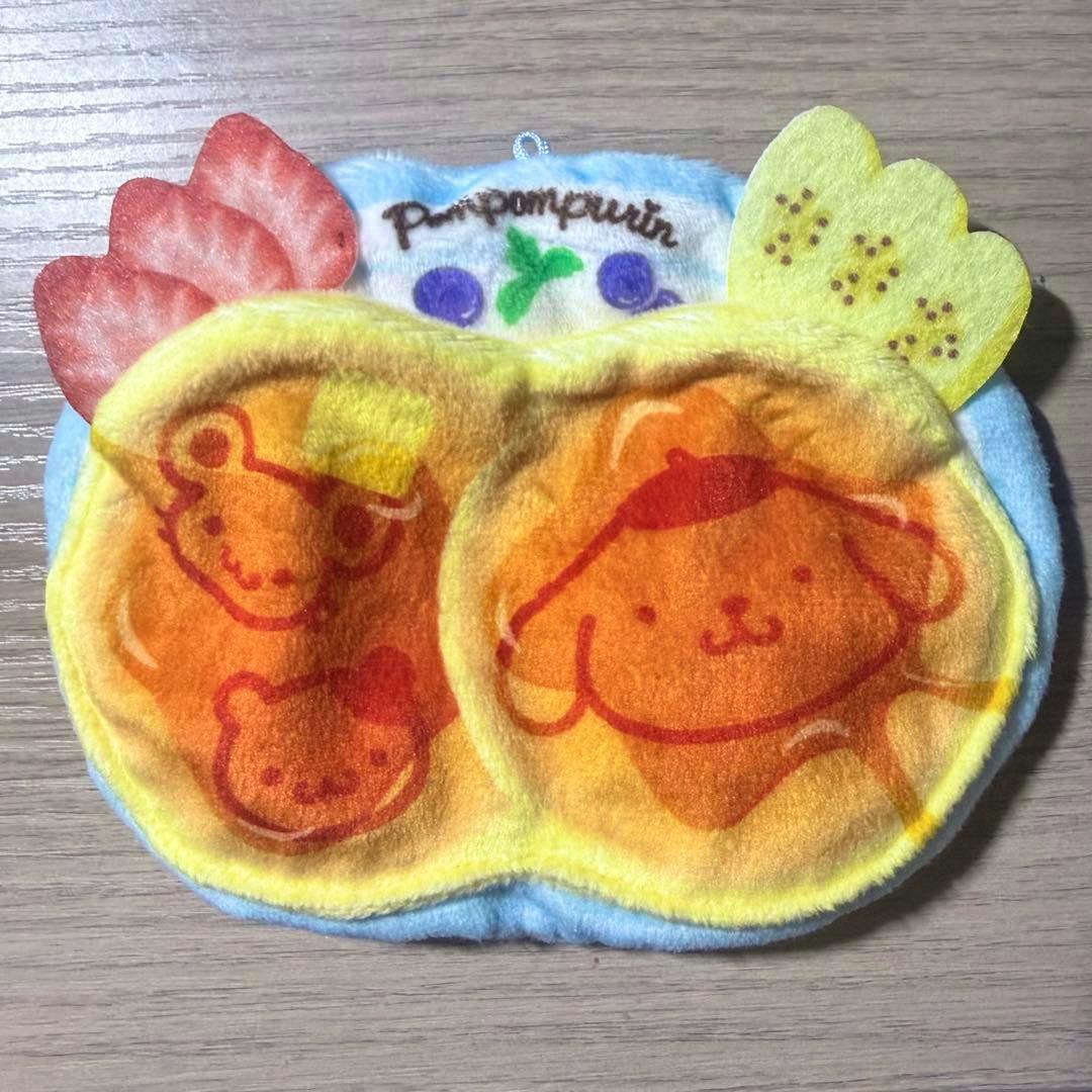 Front view of a pancake-shaped Pompompurin secret mascot pocket with printed character faces and fruit decorations.