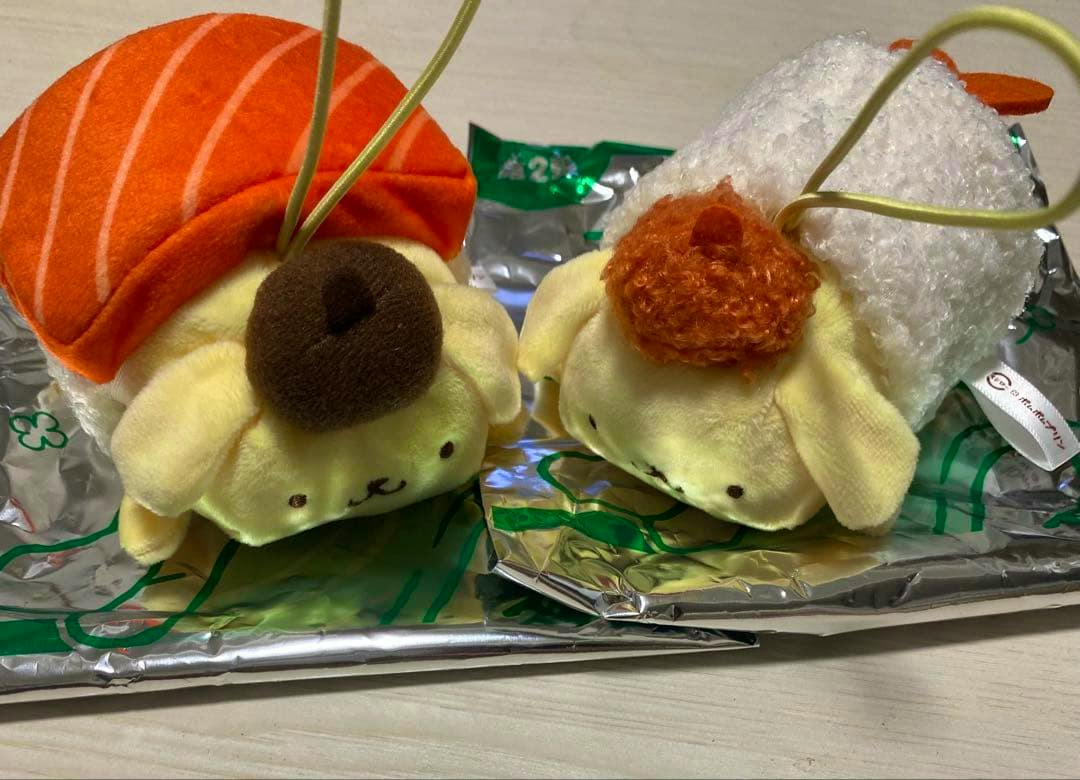 Two Pompompurin plush mascots dressed as sushi—one salmon and one shrimp—lying side by side.