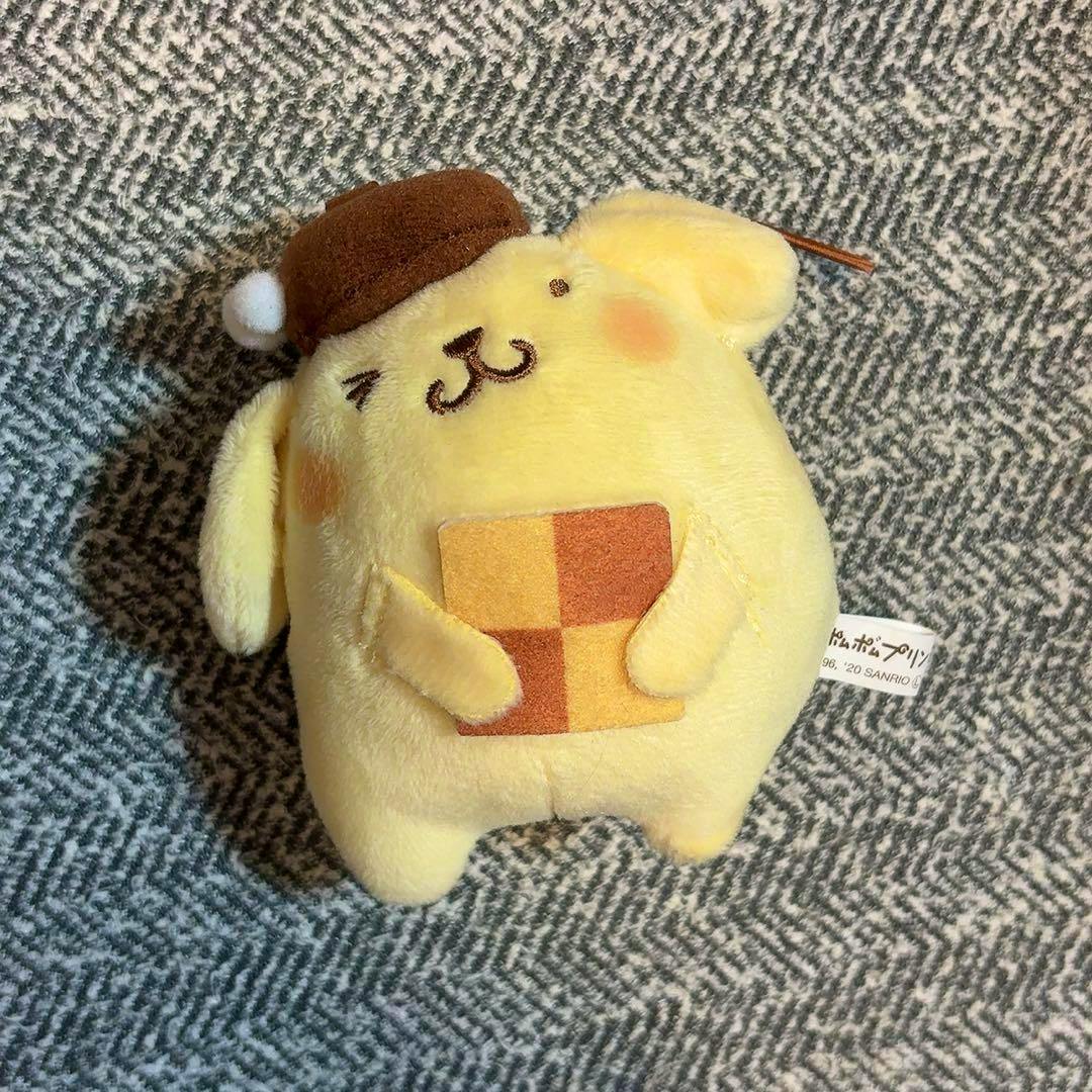 Front view of a yellow Pom Pom Purin plush mascot wearing a brown hat and holding a checkered square on patterned fabric.