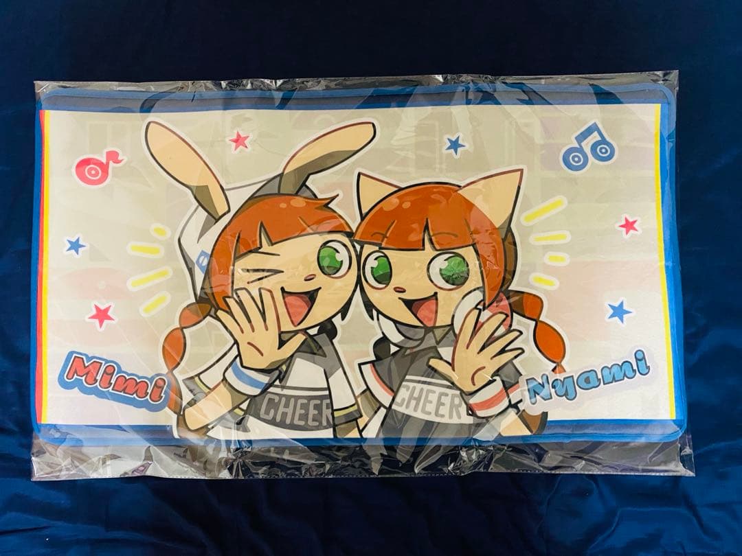 Rectangular pop'n music floor mat in clear plastic packaging featuring two cartoon characters and decorative icons, photographed on a dark blue surface.