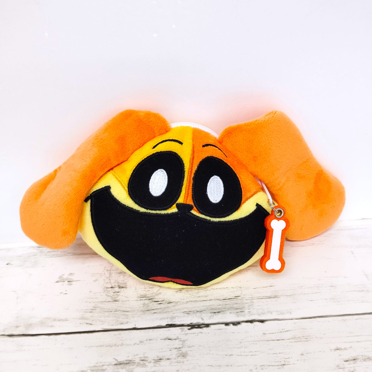 Orange Poppy Playtime dog-face polyester pouch with floppy ears and a bone-shaped zipper charm on a white wooden surface.