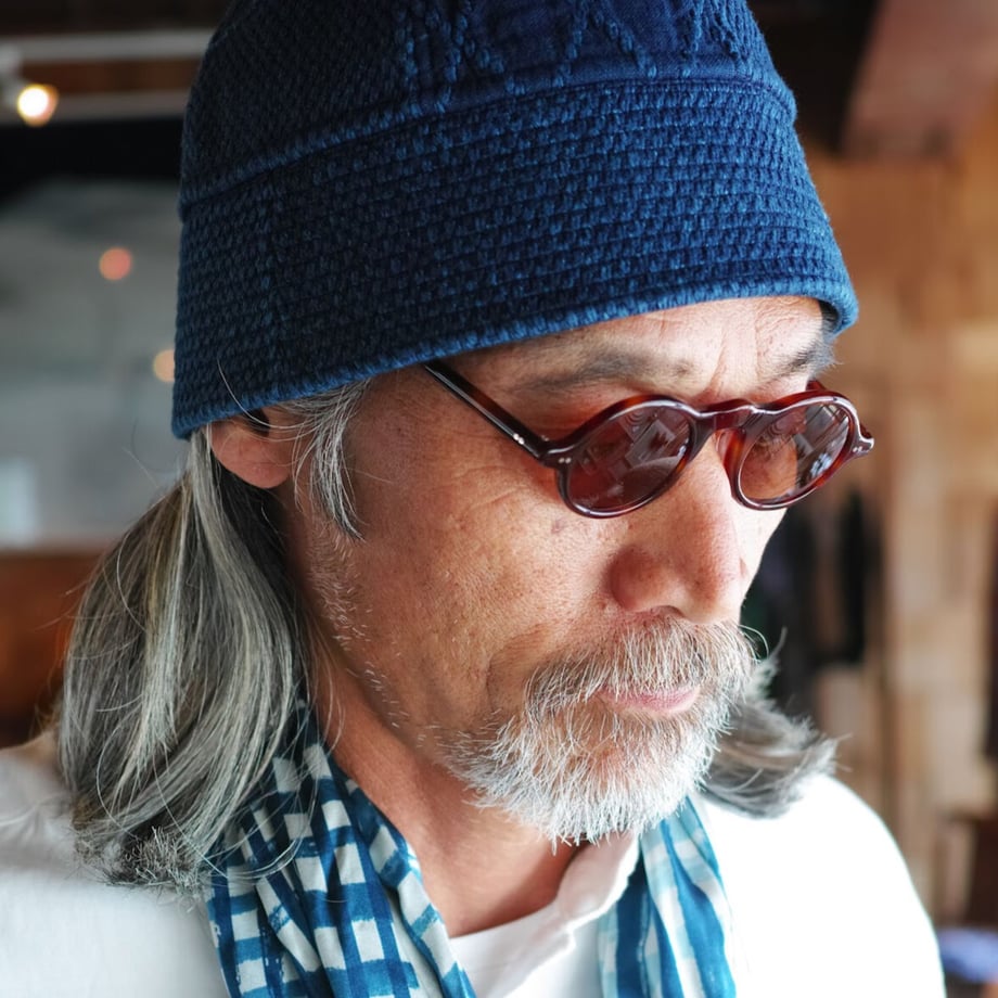 A man wearing round sunglasses, a blue knit cap, and a checked scarf, shown from the shoulders up.