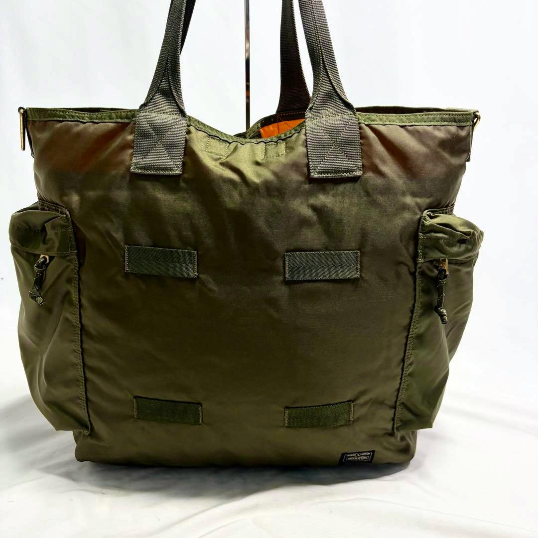Khaki nylon tote bag with top handles, two exterior side pockets, and a small logo patch at the lower right.