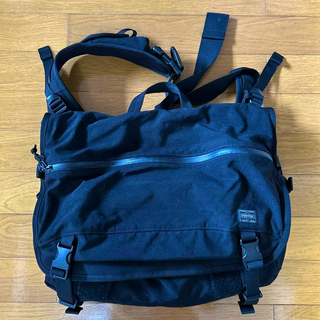 Front view of the PORTER KLUNKERZ messenger bag showing the flap with a horizontal zipper, buckle closures, and a small Porter label on a wooden floor.