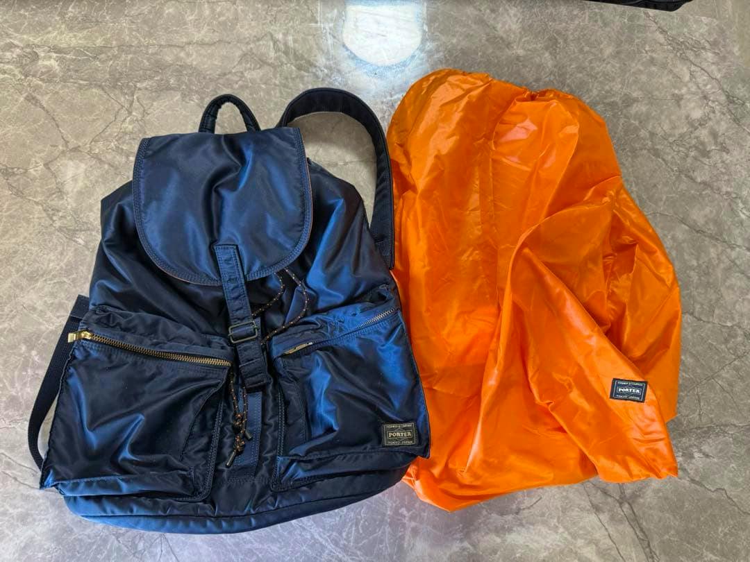 Front view of a PORTER Tanker iron-blue nylon backpack shown with an orange liner beside it on a gray surface.