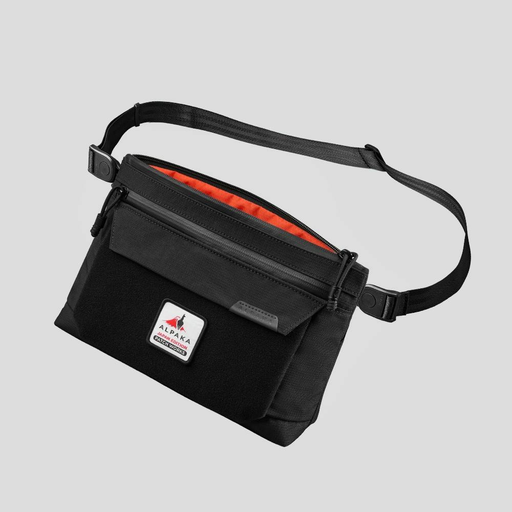 Black zip pouch with adjustable strap, front panel patch, and orange interior lining.