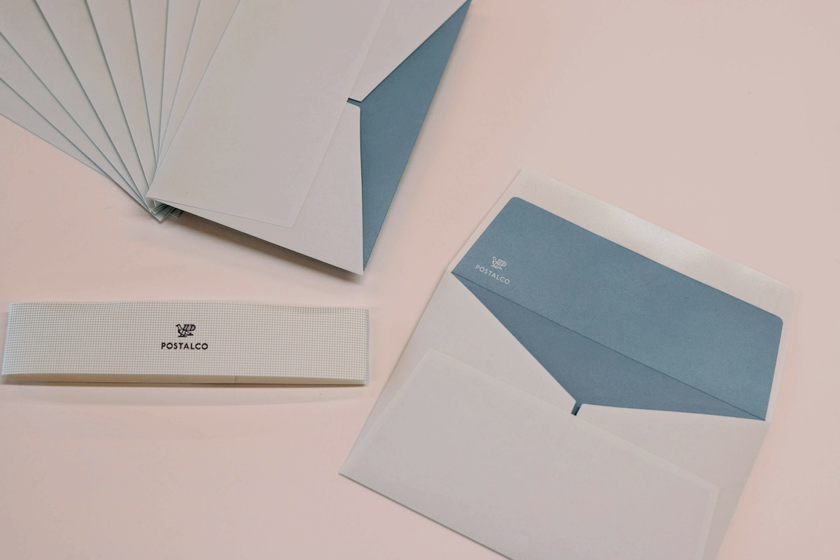 Several POSTALCO River envelopes displayed fanned and one opened to show the blue interior with a branded paper band nearby.