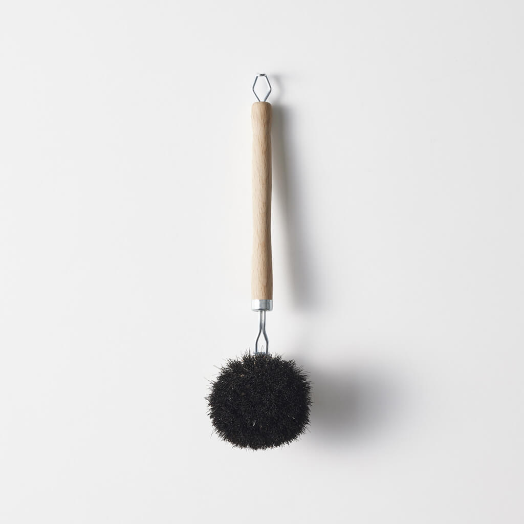 A pot and frying pan cleaning brush with a wooden handle and round black bristle head hanging against a white background.