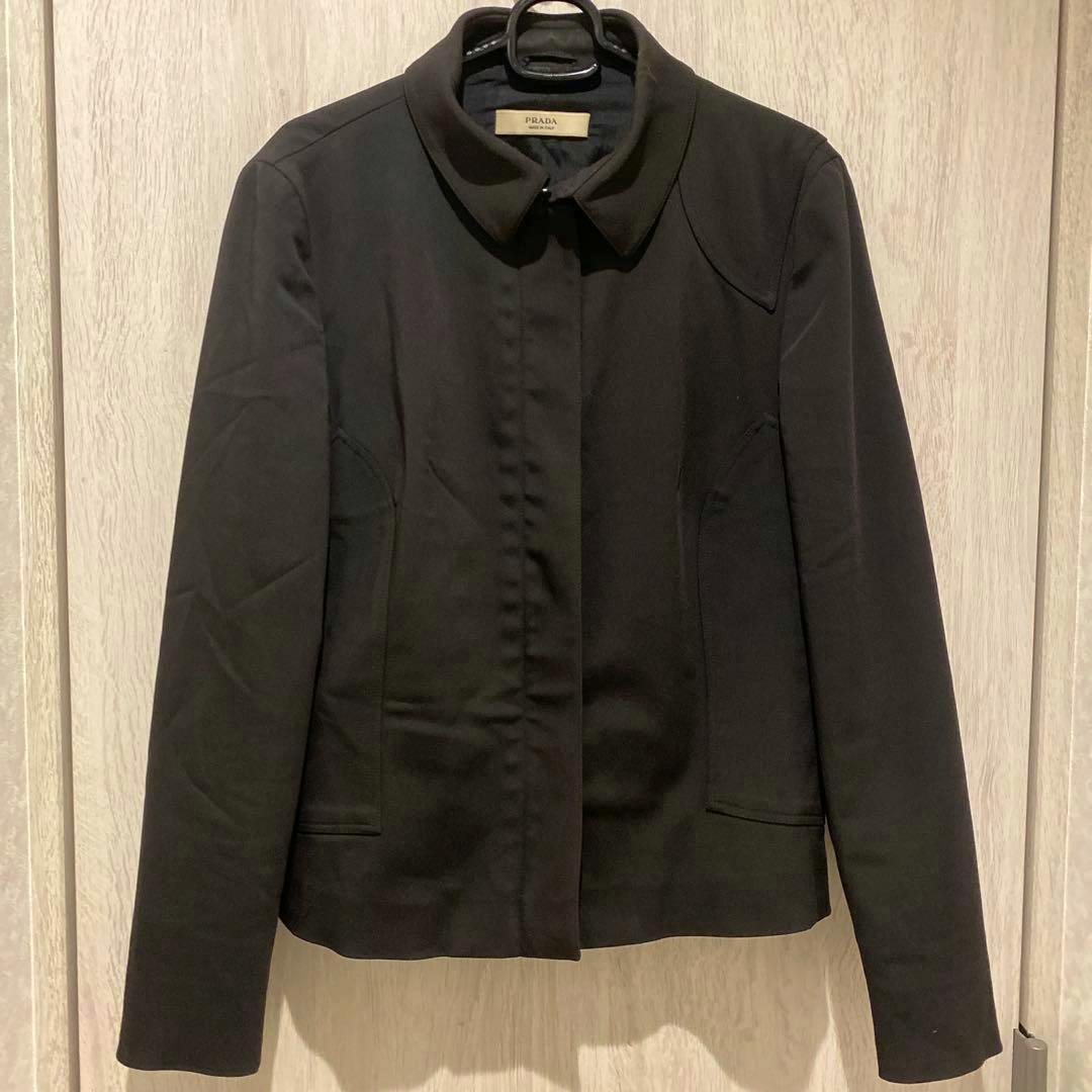 Front view of a black Prada men's nylon jacket hanging on a hanger against a wood background.