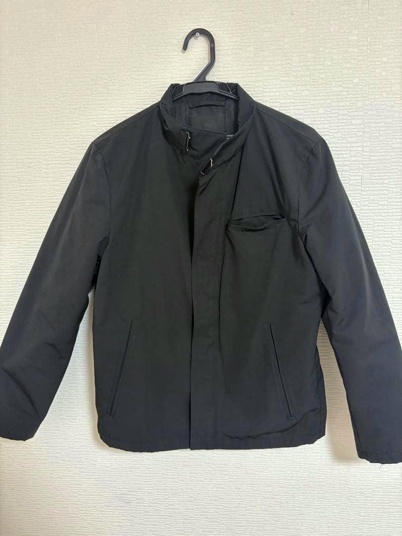 Front view of a black men's PRADA biker-style jacket hanging on a hanger against a textured wall.