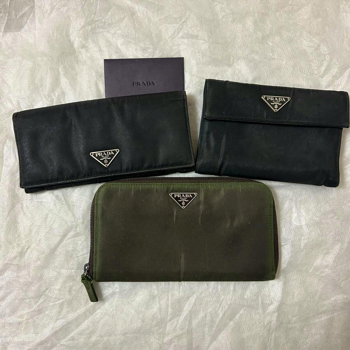 Three PRADA wallets (wallet, cow leather) with triangular logo plates and a PRADA card laid on a light background.