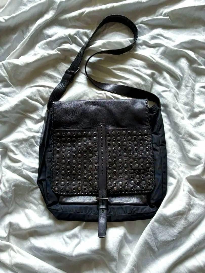 Black Prada studded leather bag laid flat on a sheet, showing the front studded panel and long adjustable strap.