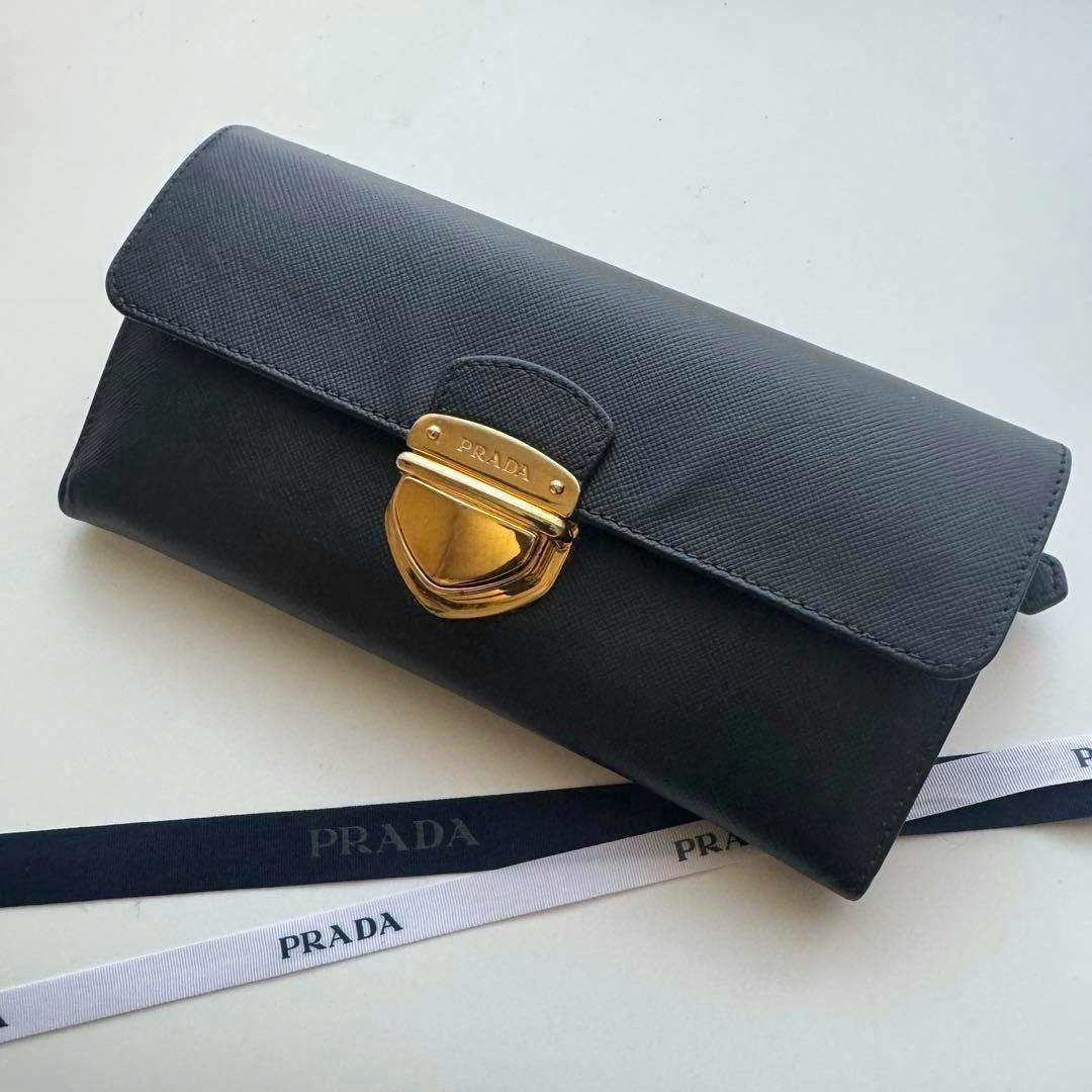 Black Prada polyester two-fold long wallet with a gold clasp on a white surface beside two PRADA ribbons.