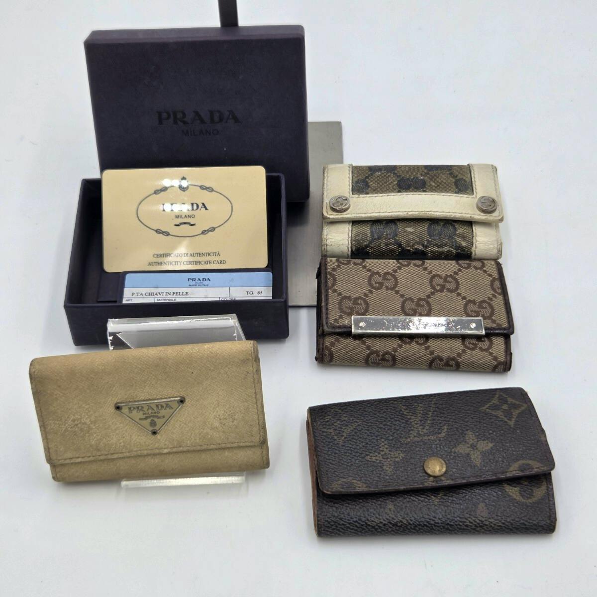 Four branded key cases, including a Louis Vuitton monogram canvas piece, a Gucci GG canvas piece, and a Prada leather case, displayed with a Prada box and authenticity cards on a white background.