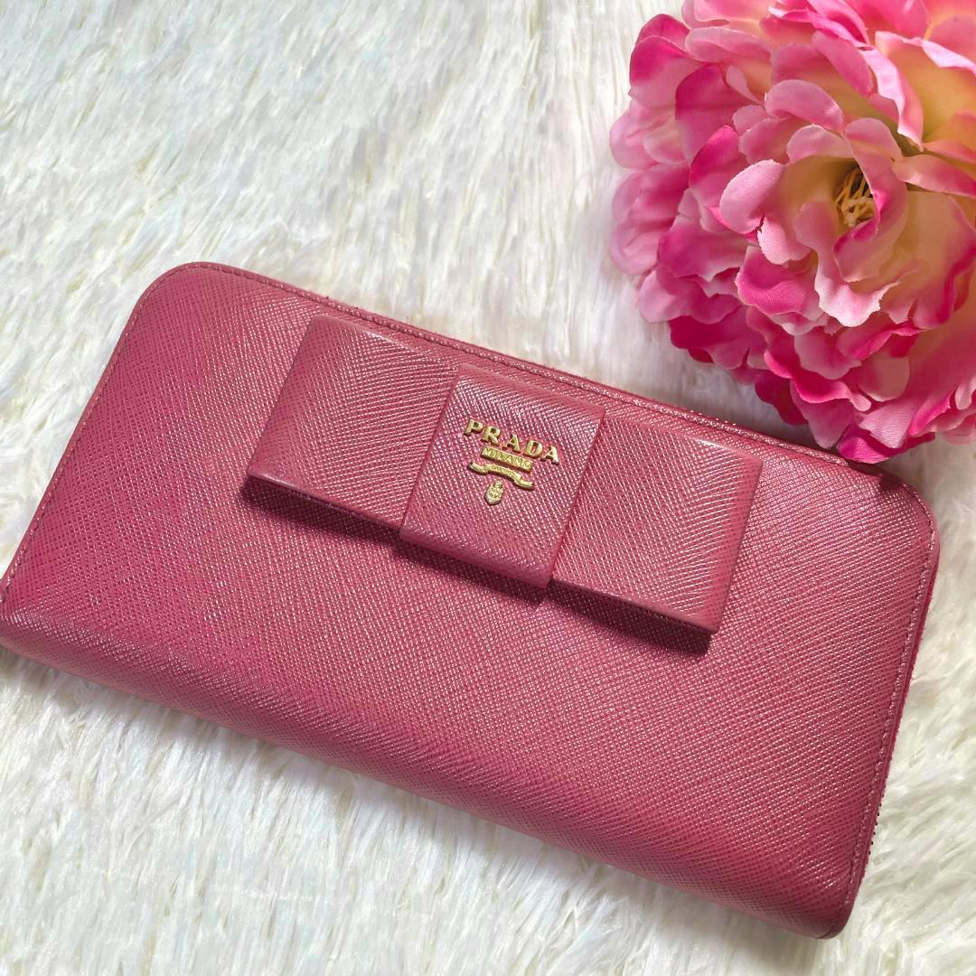 Pink saffiano leather long Prada wallet with a bow and gold Prada logo on a white fur surface beside a pink flower.