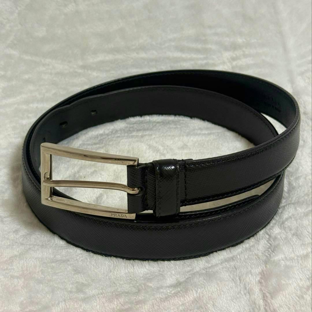 Black Prada Saffiano leather belt with a silver buckle coiled on a light fabric background.