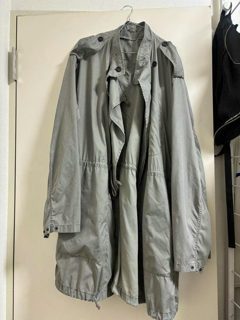 Front view of a gray men's nylon military jacket hanging on a hanger against a door.