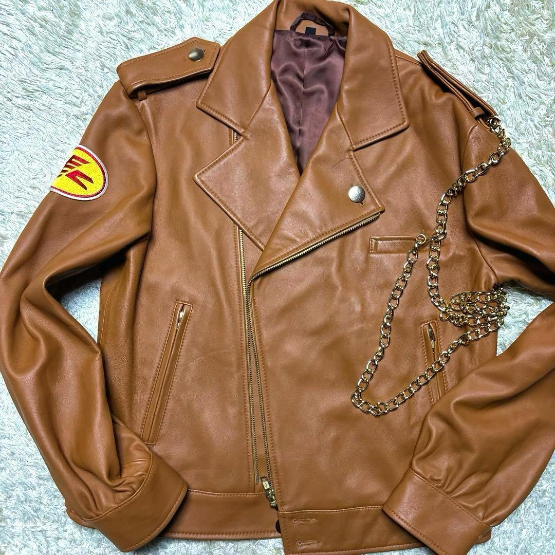 Brown lambskin double-riders jacket laid flat showing the front zipper, lapels, epaulets, a yellow/red round sleeve patch, and a gold-tone chain.